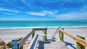 Un muelle de madera que lleva a una playa arenosa, con cielo azul despejado y mar tranquilo.