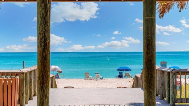 Una terraza de madera que da a una playa con agua turquesa cristalina, algunas sillas y sombrillas.