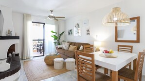 A modern living room with a white sofa, wooden dining table, and chairs. There is a ceiling fan, a woven pendant light, and a large mirror on the wall.