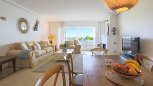 A living room with a sofa, coffee table, and dining table with chairs. There is a TV and a plant on the table. - Beachfront with large garden  - Los Monteros Palm Beach (Marbella)