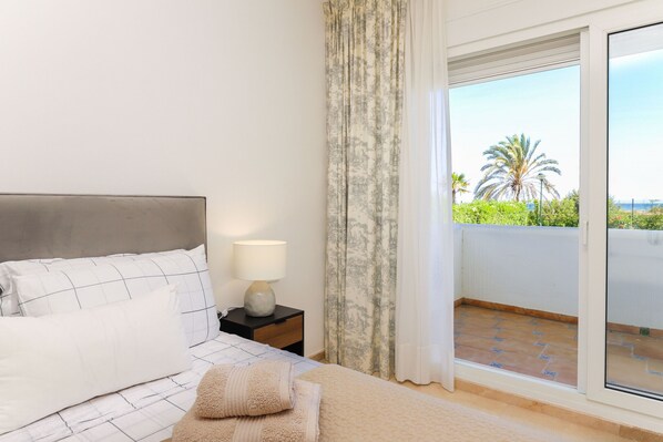 A bedroom with a bed, a nightstand, a lamp, and a view of a balcony with a palm tree and a clear sky. - Beachfront with large garden - Los Monteros Palm Beach (Marbella)
