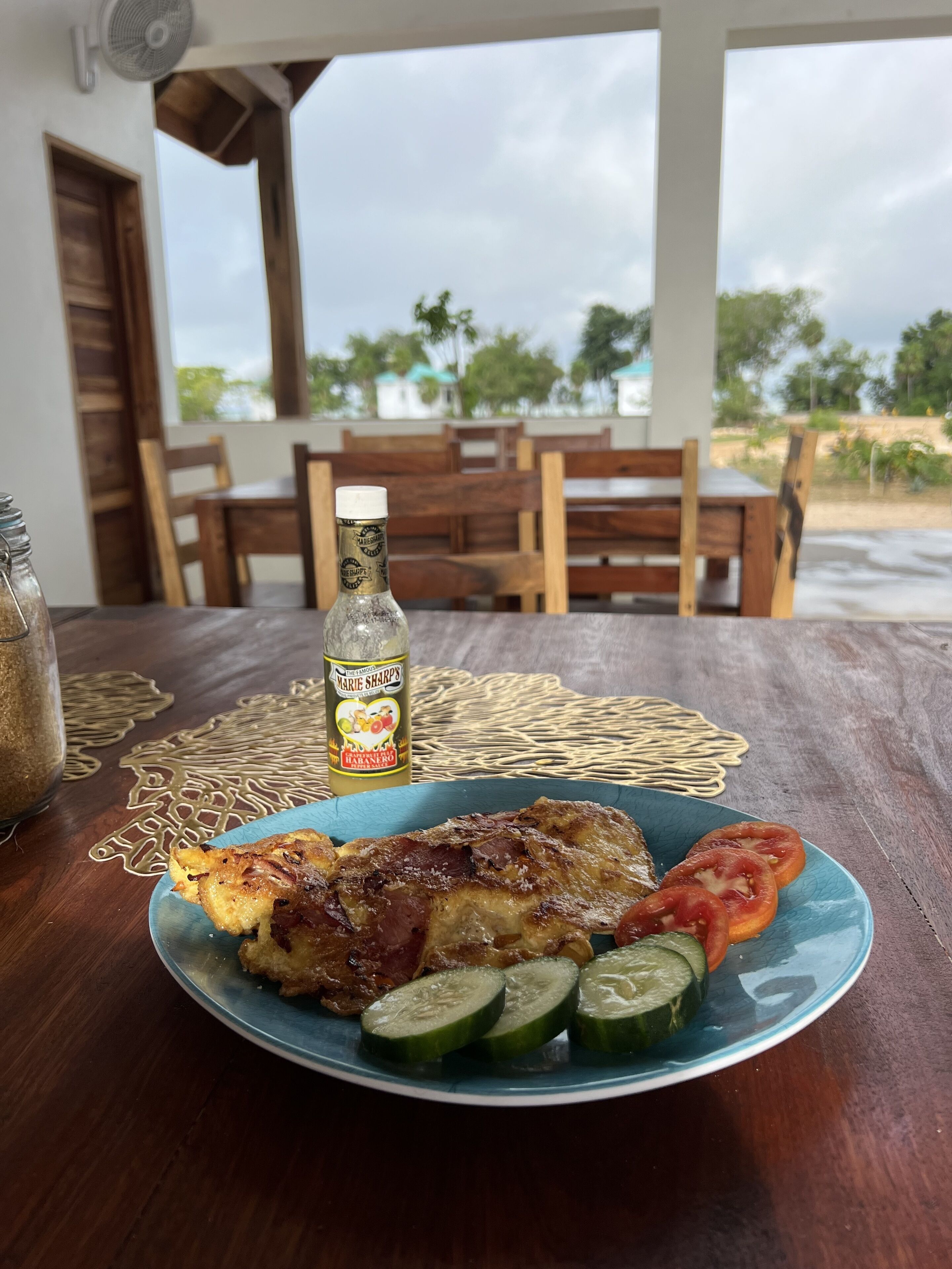 Ateria puupöydällä, jonka päällä on pullo Marie Sharpin habanero-kastiketta, lautanen grillattua lihaa, tomaatteja ja kurkkuja, ja näkymä terassille, jossa on puuhuonekaluja ja vihreää kasvillisuutta.