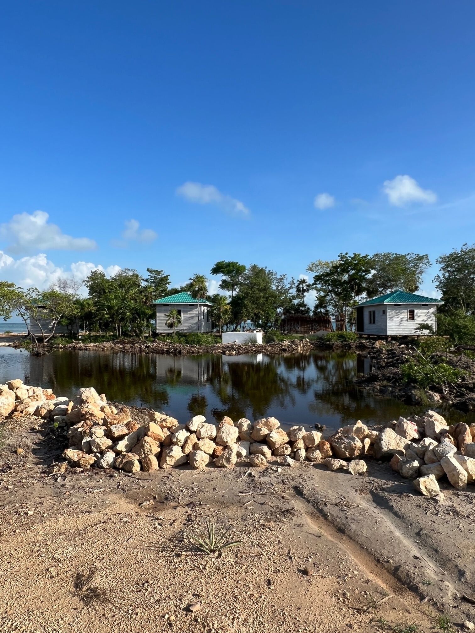 A serene lakeside setting with two small cabins, a rocky shoreline, and surrounding greenery.