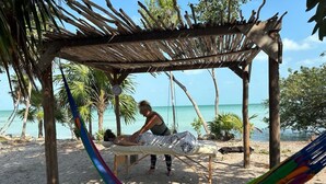 Ein Strandambiente mit einer Hängematte, einem Holz-Massagetisch und einer Person, die eine Massage erhält.