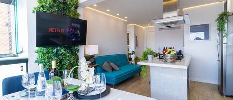 A modern living room with a teal sofa, a dining area with a white table set for two, and a kitchen with a built-in island.