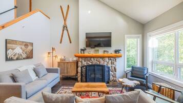 A spacious living room with a stone fireplace, a mounted TV, and a large window offering a view of trees.