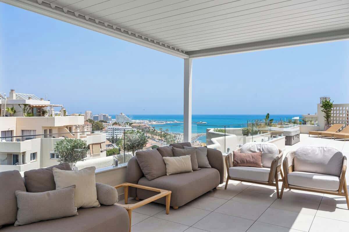 A rooftop terrace with a view of the sea, furnished with sofas and chairs, and a glass railing.