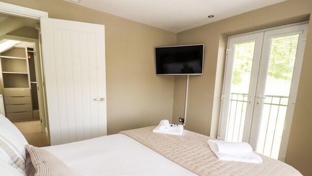 A bedroom with a bed, a TV mounted on the wall, and a balcony with a view of greenery. - Pyllau Farm (Llandudno)