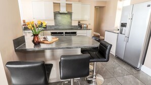 A modern kitchen with a central island, black bar stools, stainless steel appliances, and a built-in oven. - Pyllau Farm (Llandudno)