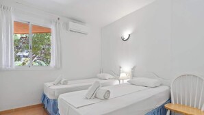 A room with two beds, a chair, a window with curtains, and a wall-mounted light.