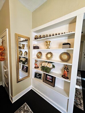 Um corredor com uma porta branca, um espelho com moldura dourada e uma estante com objetos decorativos.
