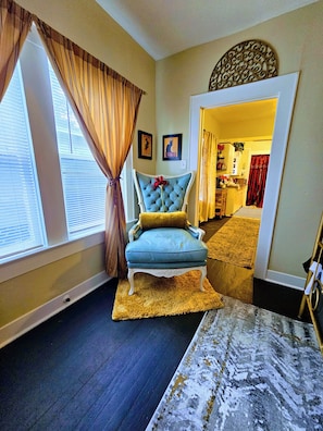 A room with a blue armchair, a patterned rug, and a view of a kitchen through an open doorway.