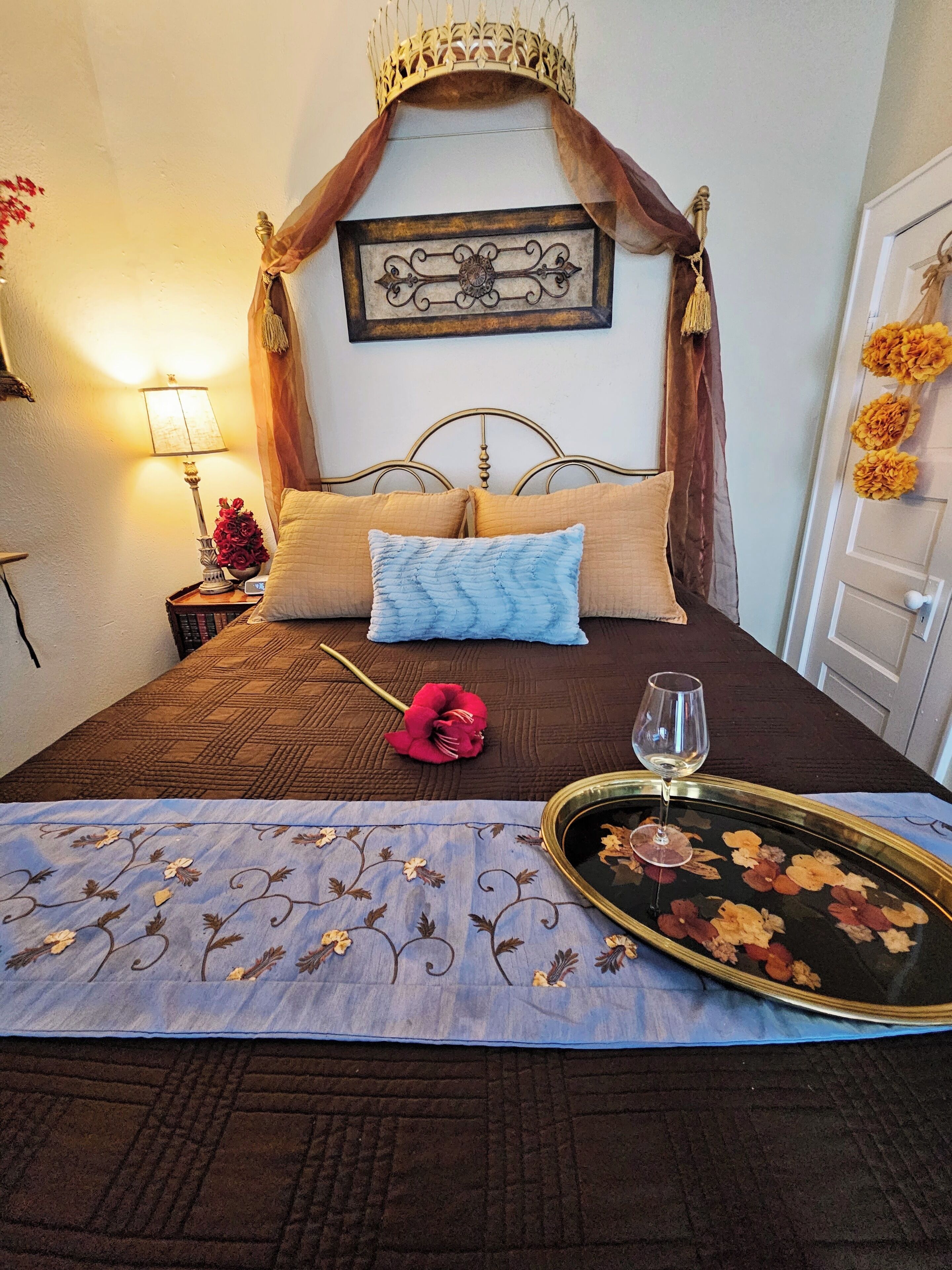 Een netjes opgemaakt bed met een decoratief hoofdeinde, een nachtkastje met een lamp en bloemen, en een dienblad met een wijnglas.