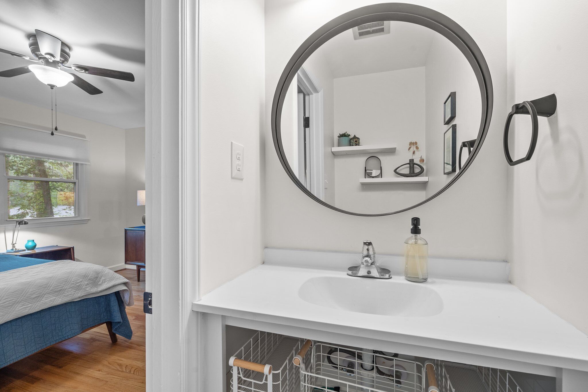 A bedroom with a bed, a ceiling fan, and a window. The bathroom features a white sink, a round mirror, and a shelf with toiletries.