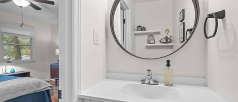 A bedroom with a bed, a ceiling fan, and a window. The bathroom features a white sink, a round mirror, and a shelf with toiletries.