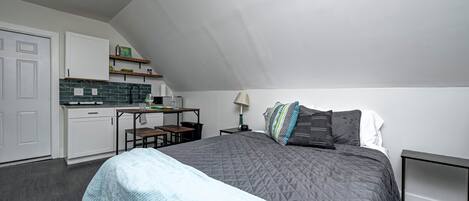 A bedroom with a bed, a desk, and a kitchenette.