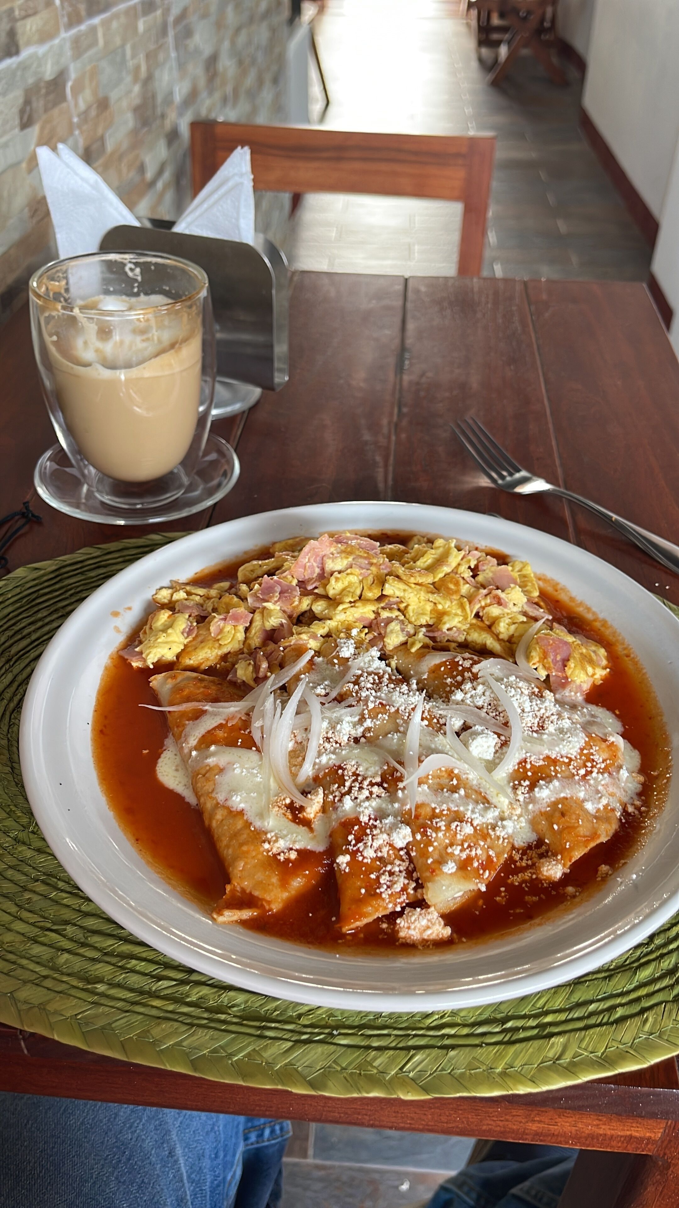 A plate of enchiladas topped with cheese, onions, and a red sauce, served with a glass of iced coffee on a wooden table.