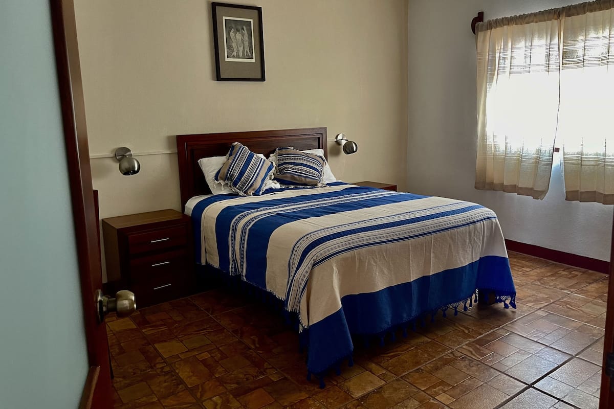 A bedroom with a bed, a nightstand, a wall-mounted lamp, a framed picture, and a window with curtains.