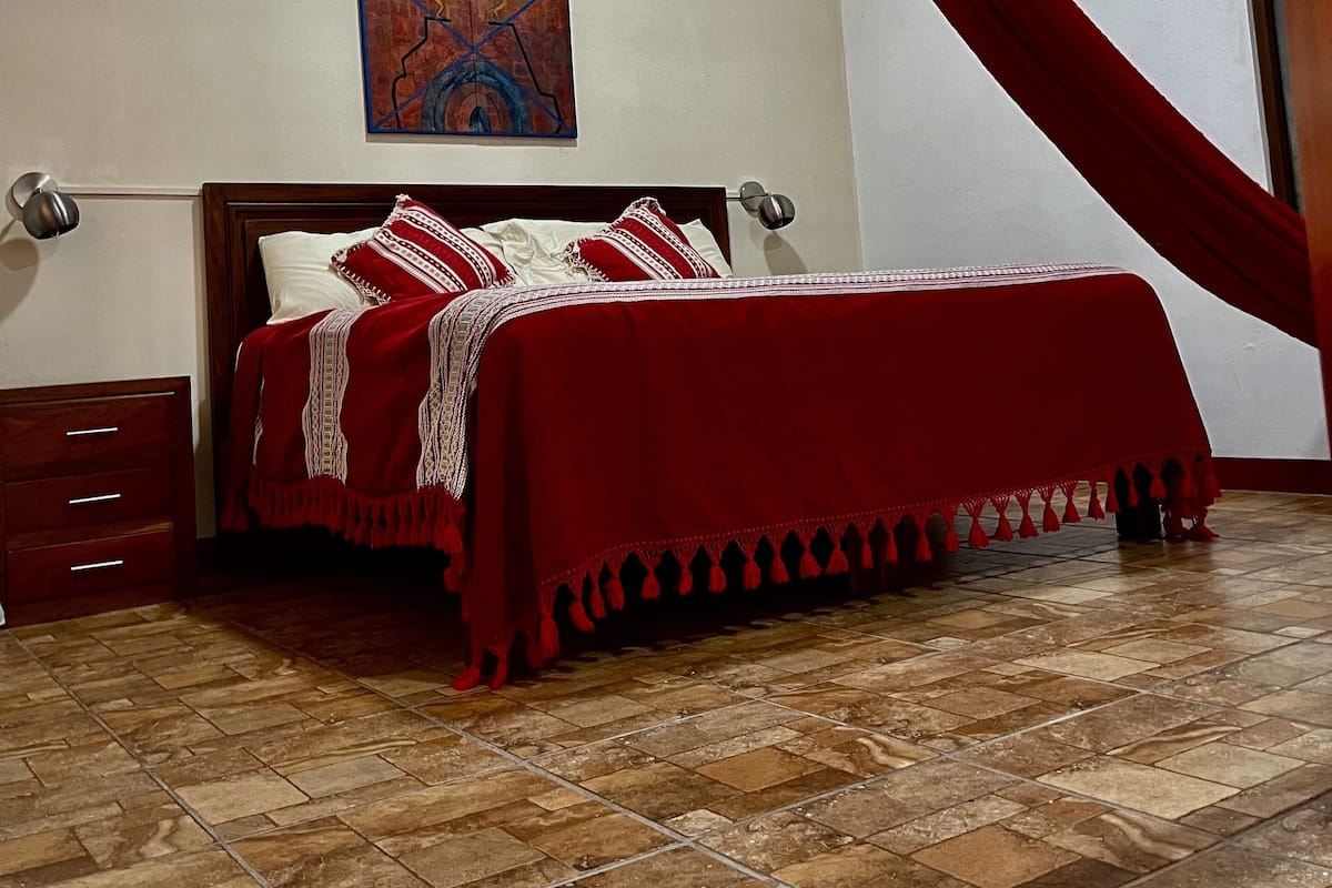 A bedroom with a bed, a nightstand, a lamp, a painting on the wall, and a red curtain.
