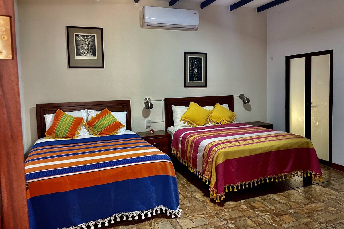 Two beds with colorful bedding, a wall-mounted air conditioner, and framed artwork on the walls.