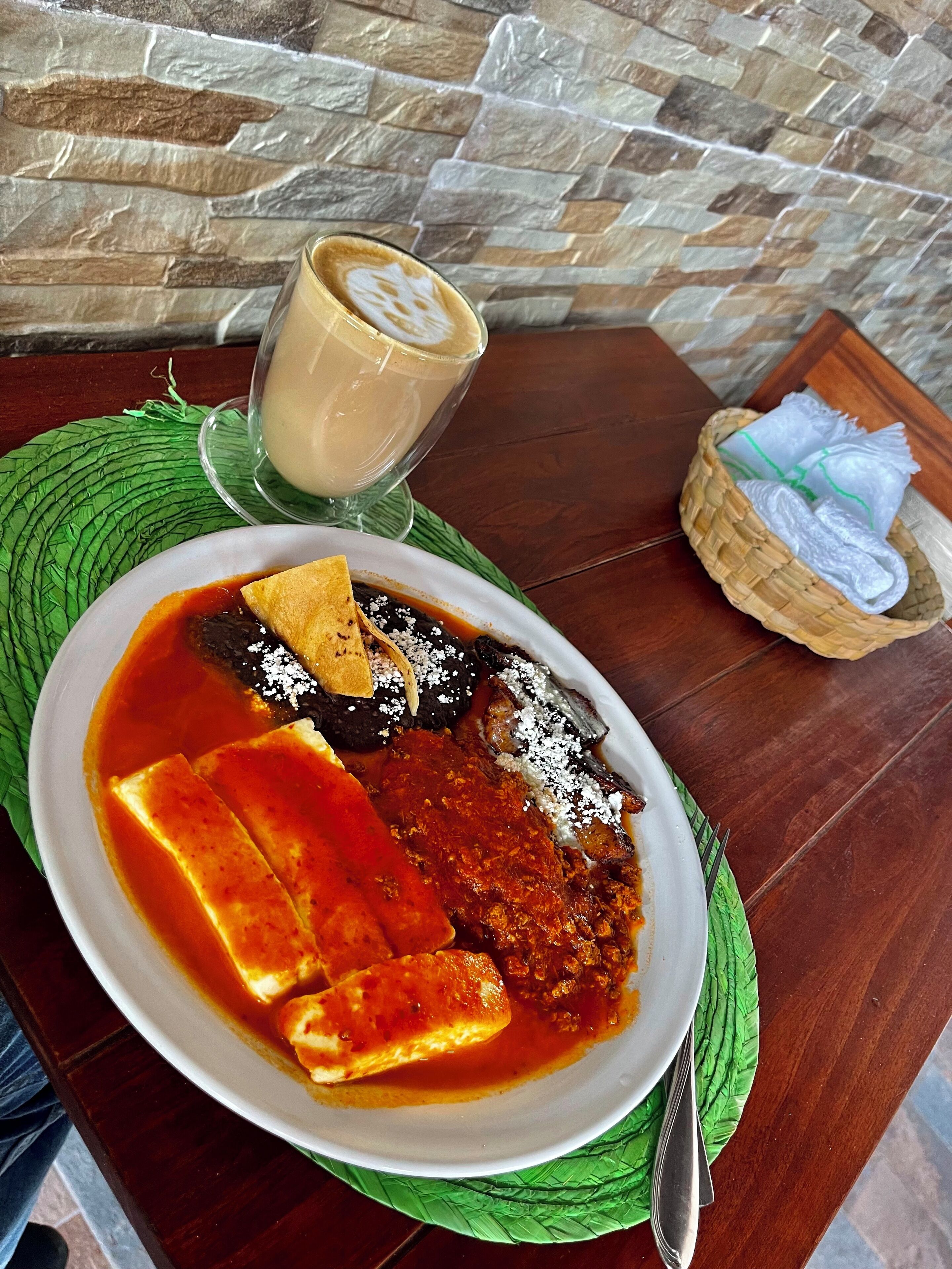 Un plato de enchiladas con salsa, una taza de café y un cesto de servilletas sobre una mesa de madera.