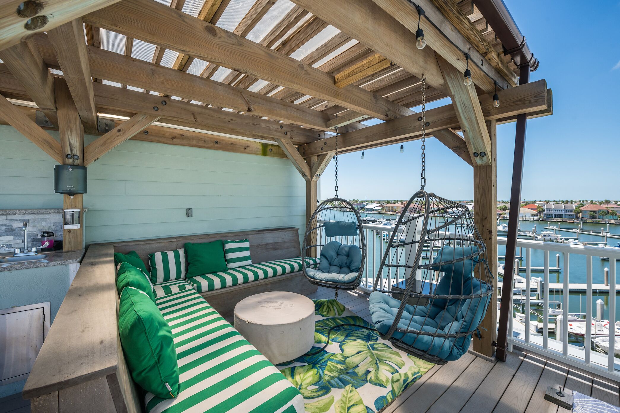 Uma área de estar ao ar livre coberta com vista para uma marina, com pérgola de madeira, almofadas listradas, uma cadeira suspensa e uma mesa redonda.