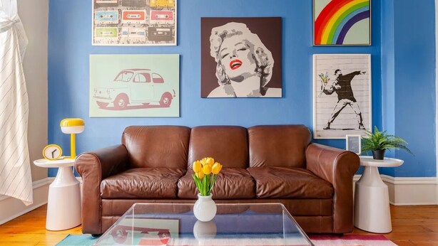 A living room with a brown leather sofa, a glass coffee table with a vase of yellow flowers, and various wall art including a car, a woman, and a rainbow. - FESTIVE GRAFITTI POP-ART 3BR - Wicker Park - Ukrainian Village (Chicago)