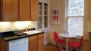 A compact kitchen with wooden cabinets, a dishwasher, a window with blinds, and a small dining area with two red chairs. - FESTIVE GRAFITTI POP-ART 3BR - Wicker Park - Ukrainian Village (Chicago)