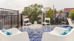 Une terrasse sur le toit, dotée de mobilier d’extérieur blanc, d’un tapis à motifs et offrant une vue sur les bâtiments et les arbres.