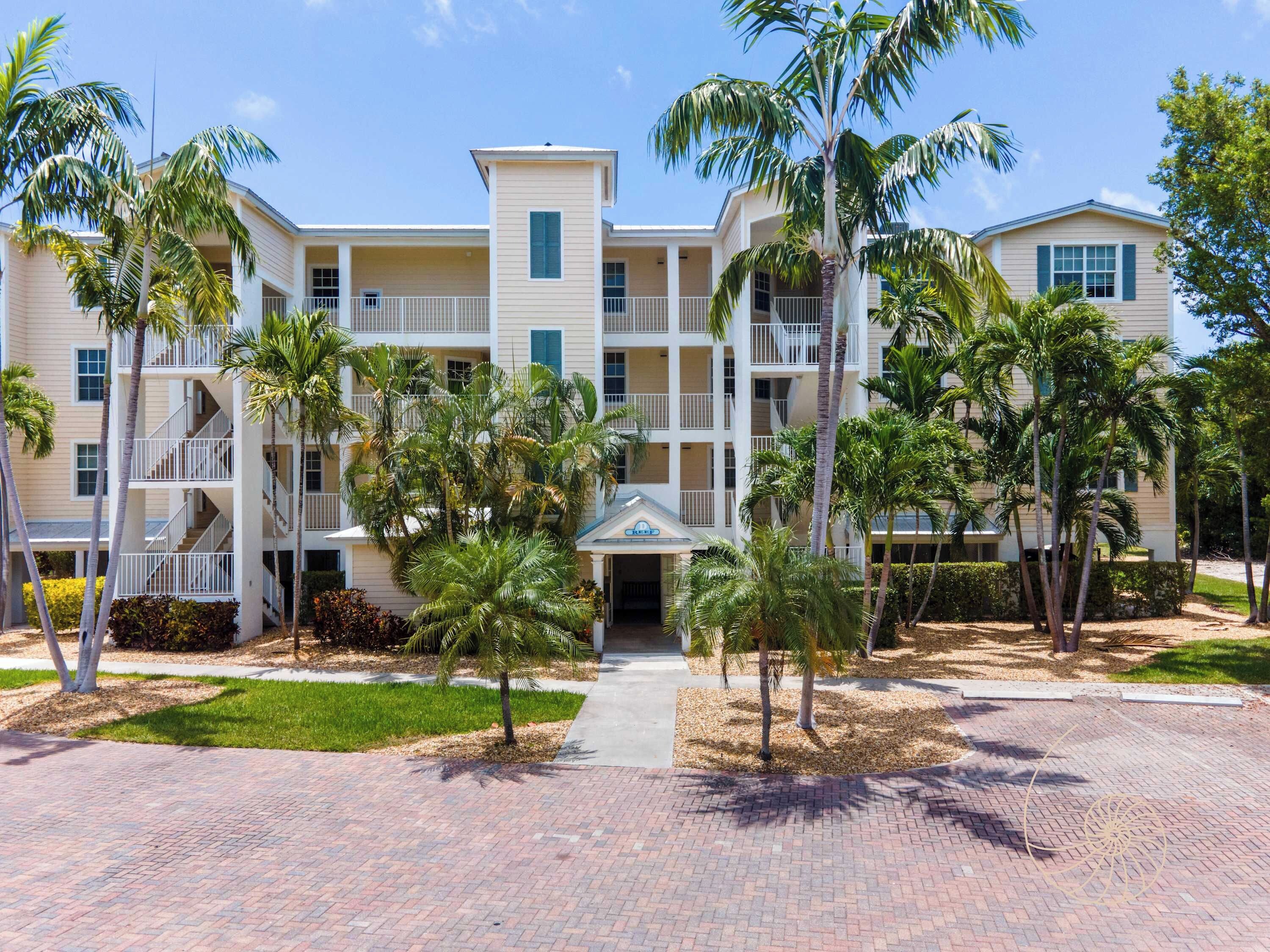 A multi-story hotel with a central entrance, surrounded by palm trees and a paved walkway.