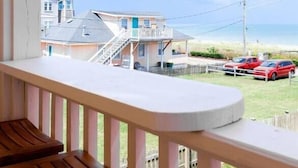 Ein Balkon mit Holztisch, Blick auf einen Strand mit Gebäuden und parkenden Autos.