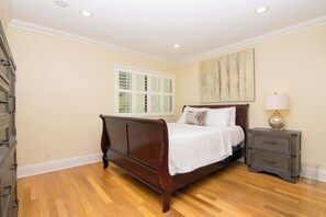 Un dormitorio con cama de madera, una mesita de noche con lámpara, una ventana con persianas y un cuadro en la pared.