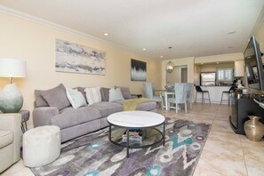A living room with a sofa, ottoman, coffee table, and a rug. There is a kitchen area with a refrigerator, stove, and chairs. - First Floor 2bd/2ba Fully Remodeled Condo in Saddlebrook (Wesley Chapel)