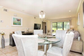 A dining area with a glass table, white chairs, a chandelier, and a painting on the wall.
