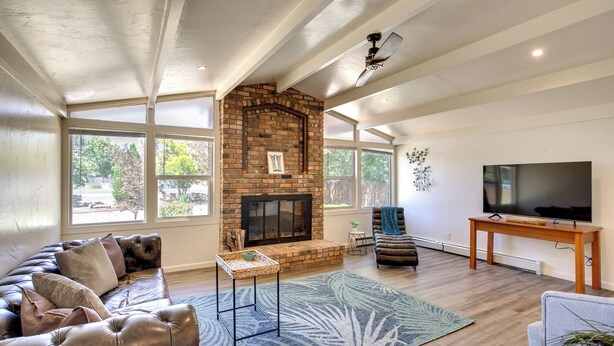 A spacious living room with a fireplace, large windows, a leather sofa, a television, and a ceiling fan. - Grand Junction Vacation Rental w/ Patio & Grill! (Grand Junction)