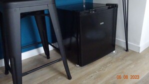 Un petit réfrigérateur noir avec le logo de la marque bien visible, un tabouret noir et un mur bleu en arrière-plan.