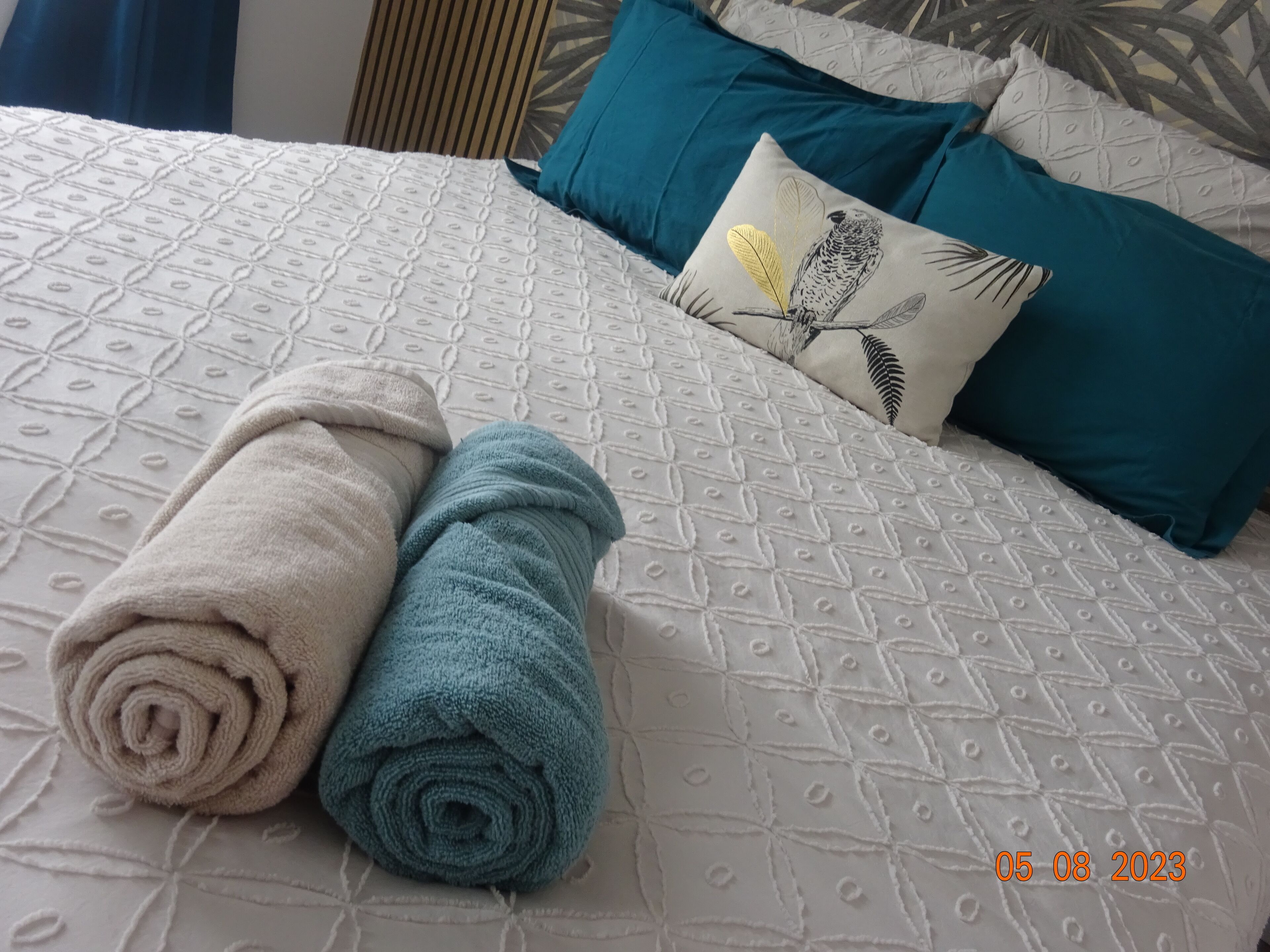 A bed with a patterned white duvet, two rolled towels in beige and teal, and a pillow with a bird design.