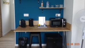 Une petite cuisine équipée d’un four à micro-ondes, d’une machine à café et d’un petit réfrigérateur. On y trouve également deux tabourets, une poubelle et un ventilateur.