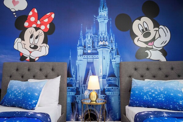 Ein Zimmer mit zwei Betten, jedes mit einem Mickey Mouse Kopfkissen, einer Lampe auf einem Nachttisch und einer Wandmalerei mit einem Schloss.