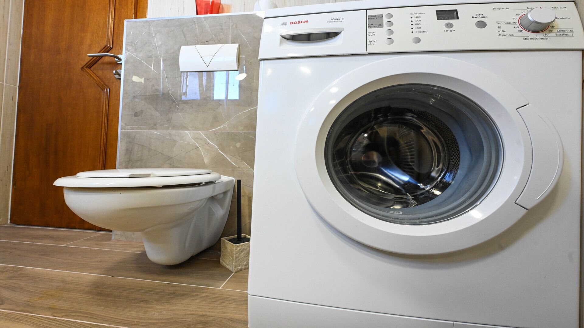 A Bosch washing machine in a bathroom with a toilet and a marble-tiled wall.