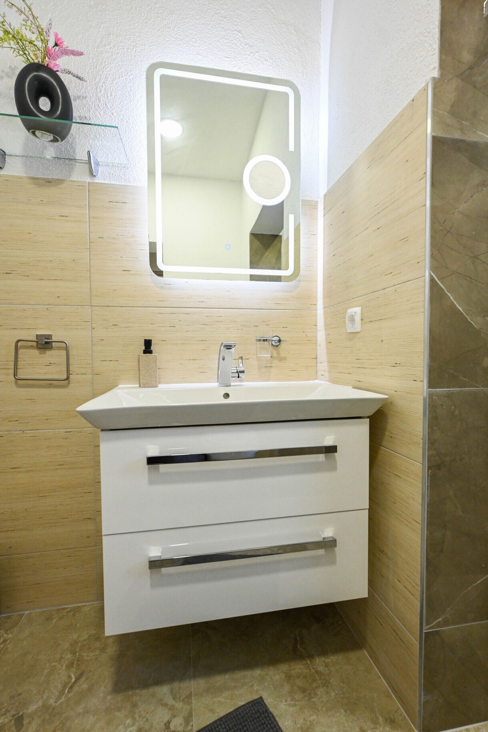 Een moderne badkamer met een wit wastafelmeubel, een spiegel met ingebouwde verlichting, een handdoekhouder en een douchehoek.