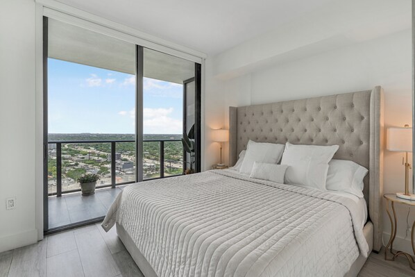 A bedroom with a large window, a tufted headboard, and a view of a cityscape.