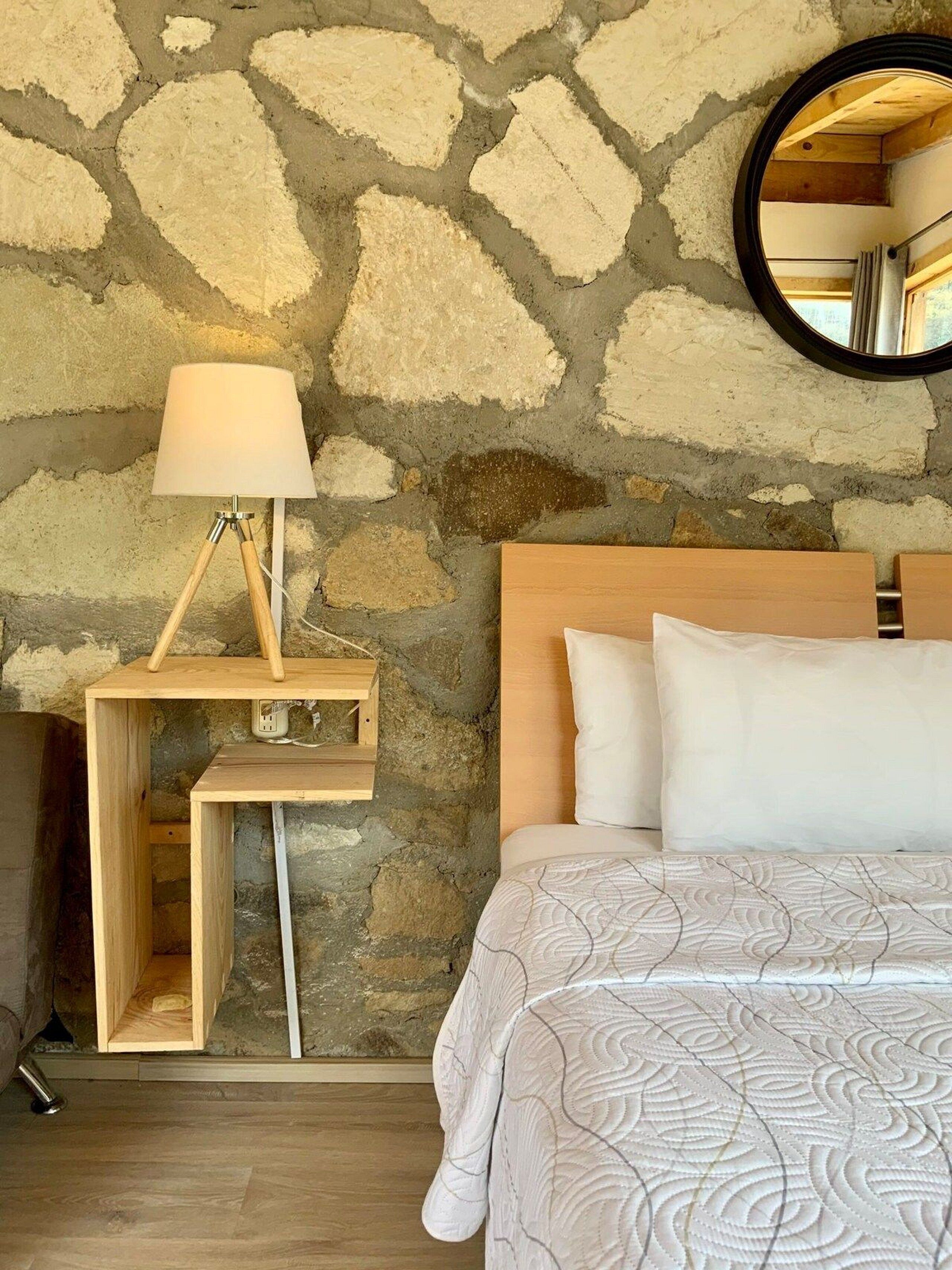 Une chambre avec un mur en pierre, une table de chevet en bois avec une lampe, un lit avec une literie blanche et un miroir rond accroché au mur.