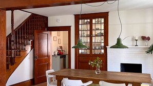 A dining area with a wooden table and white chairs, a staircase, and a kitchen area with a white brick fireplace. - At Leo's by Maison Dudley (Lac-Mégantic)