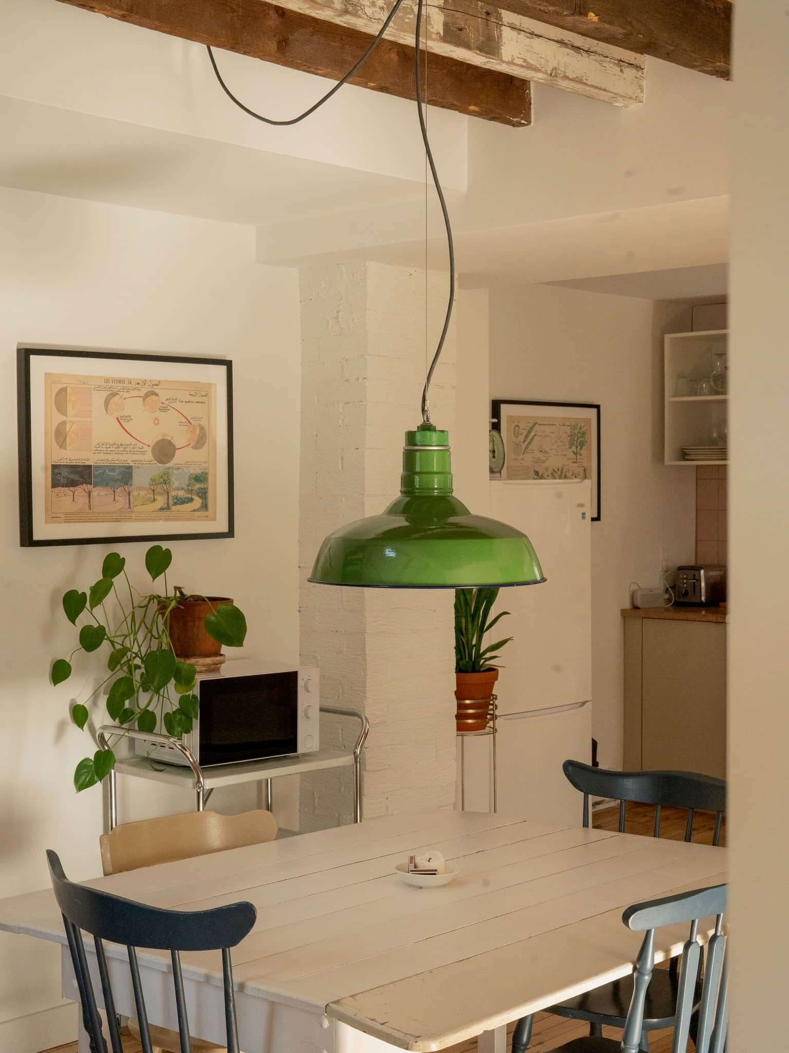 Een eetruimte met een witte tafel, blauwe stoelen, een groene hanglamp en een laptop op een bijzettafeltje.
