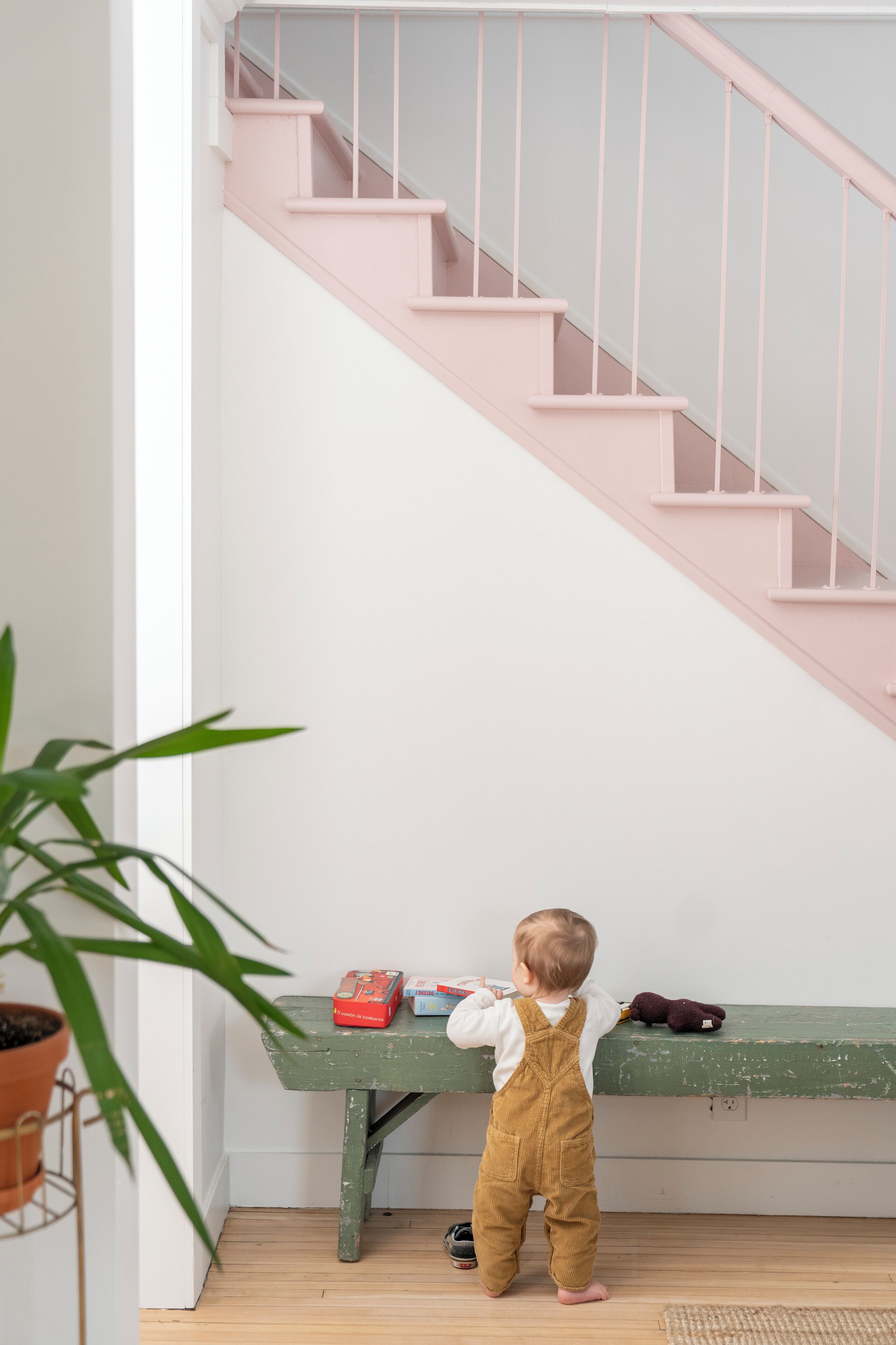 A child standing at the bottom of a staircase with a green bench and a potted plant nearby.