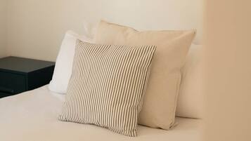 Un letto con due cuscini, uno a righe e uno tinta unita, in una stanza con una parete spoglia e un comodino scuro.