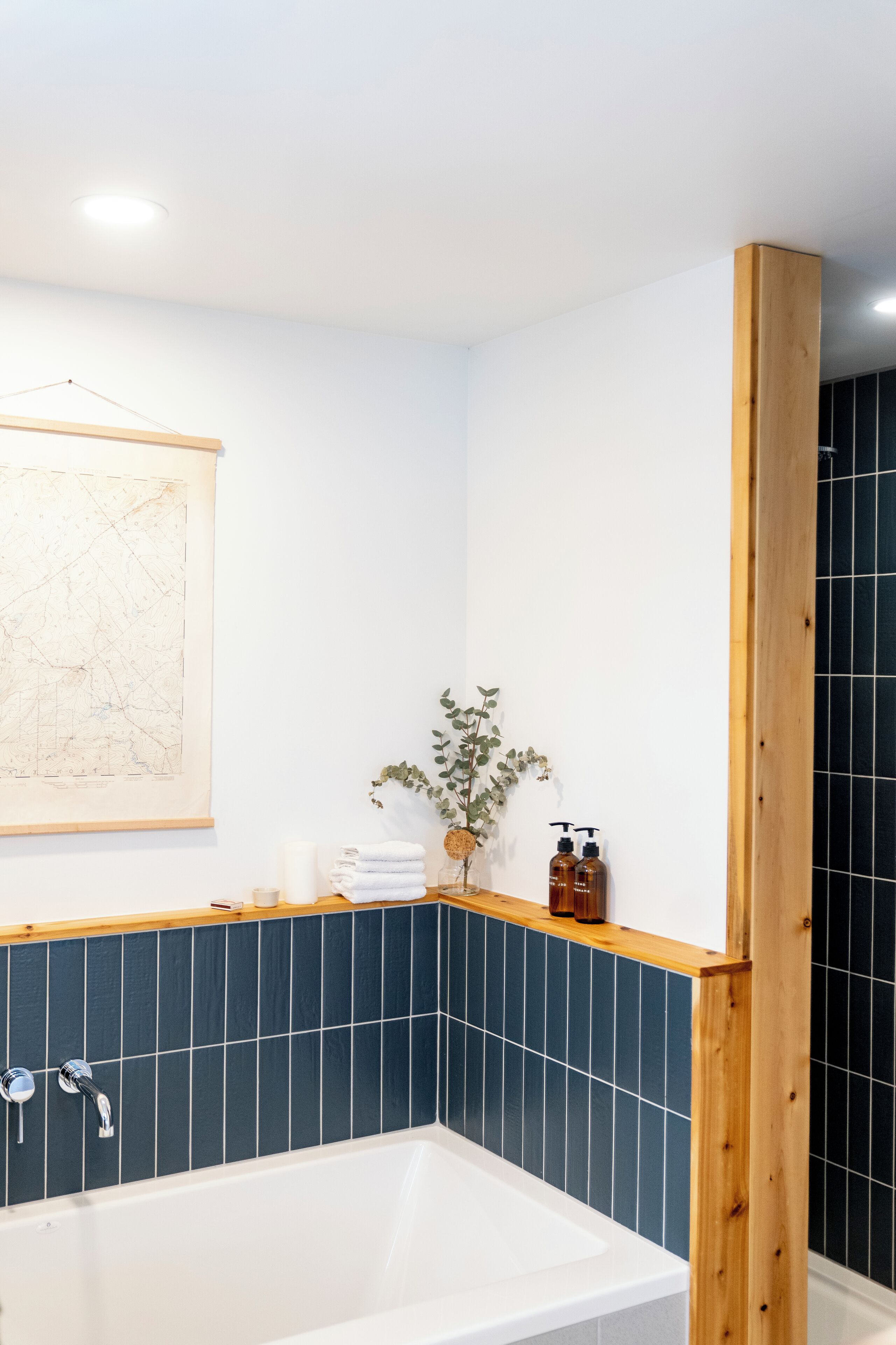 Ein modernes Badezimmer mit einer weißen Badewanne, dunkelblauen Fliesen, einem Holzbalken, einer Karte an der Wand und Pflegeprodukten auf einem Regal.