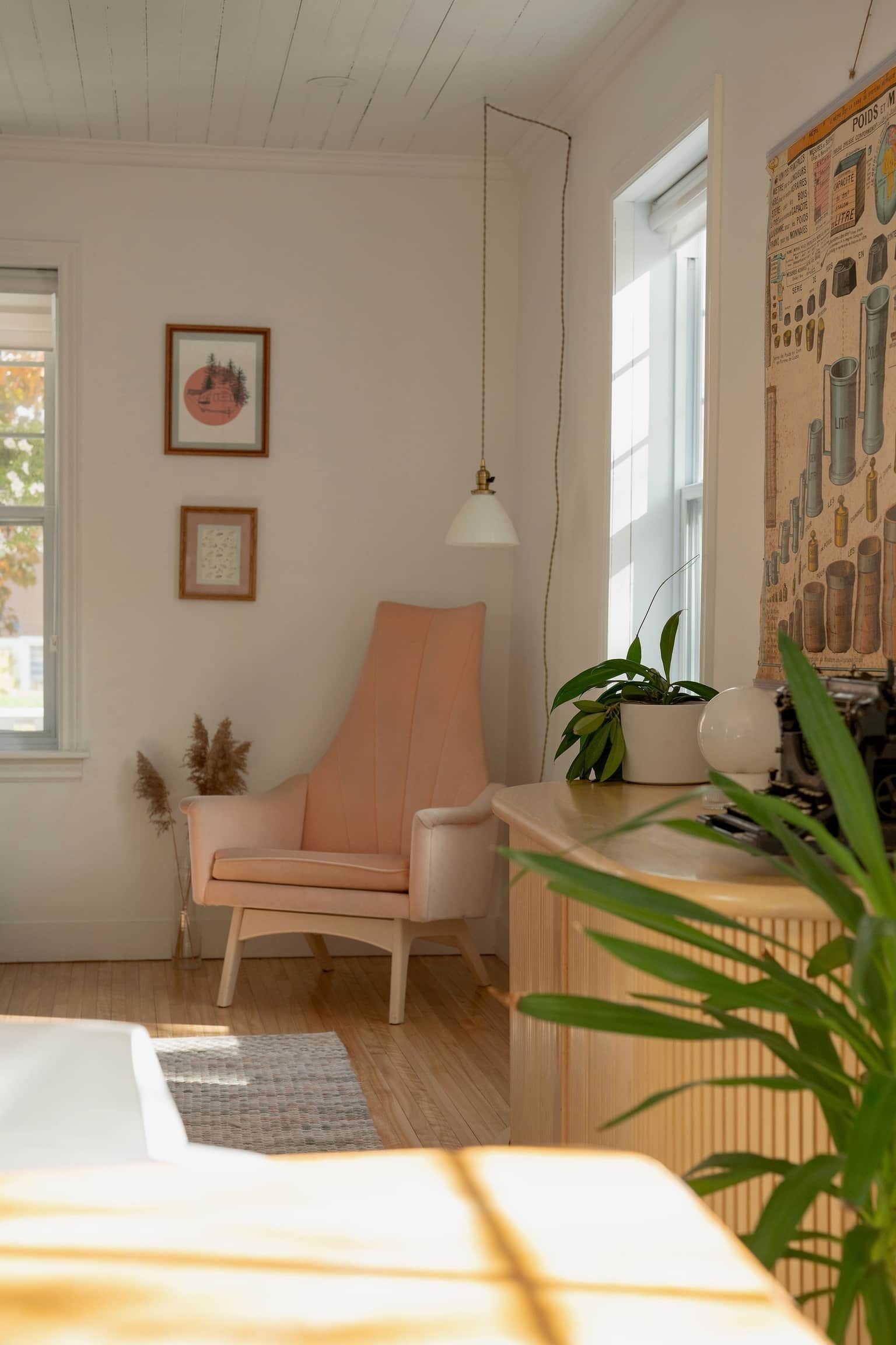 A cozy room with a wooden ceiling, a white chair, a desk with a plant, and a window with a view.
