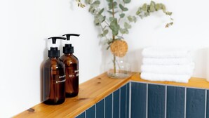 A bathroom with a wooden countertop, two brown bottles, a glass vase with a cork, and a stack of white towels. - The Boulevard by Dudley House (Lac-Mégantic)